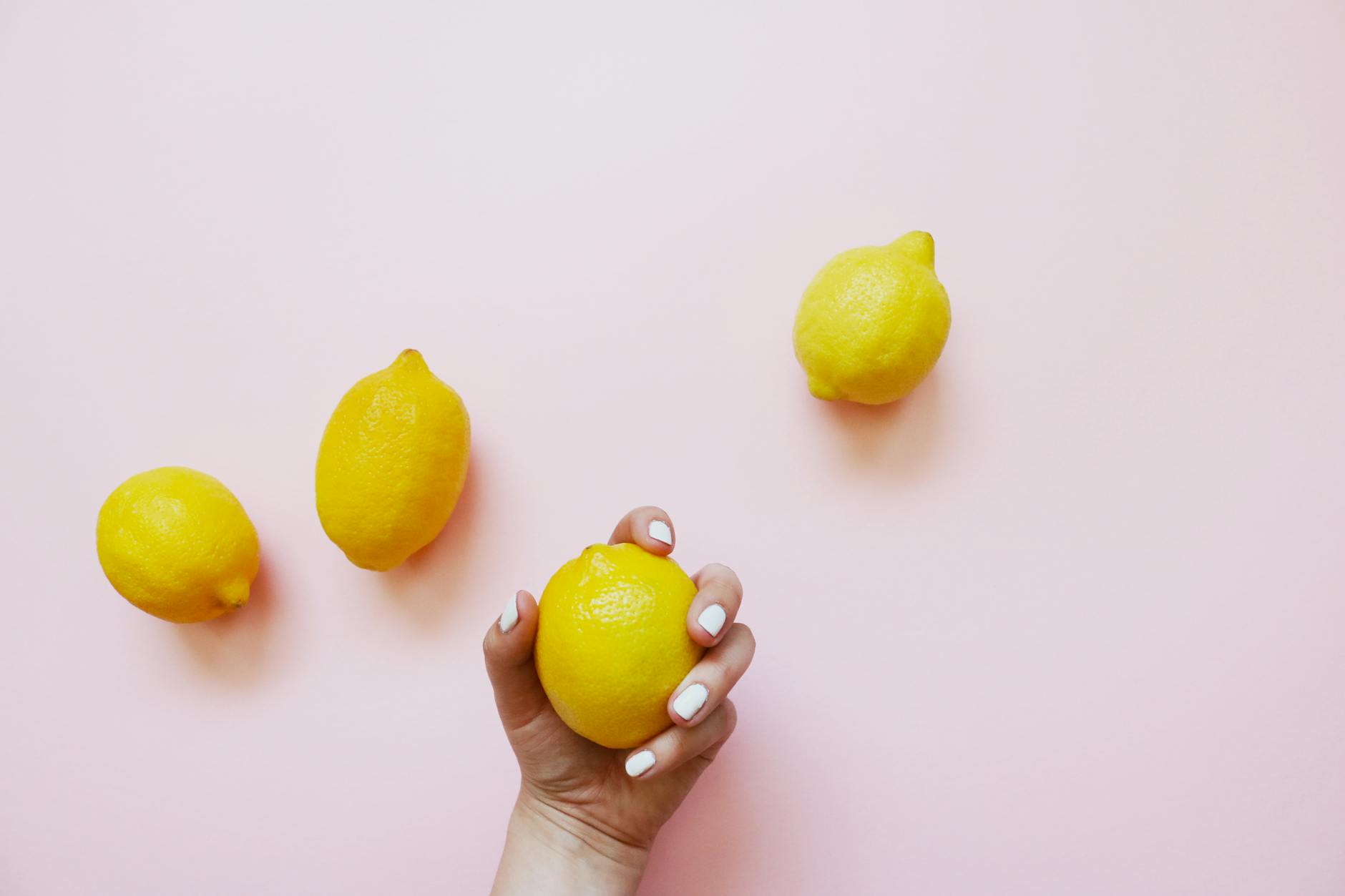 Una mano che tiene un limone su uno sfondo rosa morbido, circondata da altri limoni